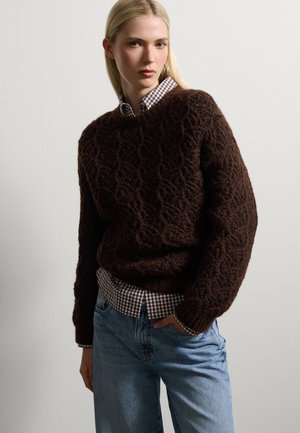 Pull en tricot marron avec un motif texturé, superposé à une chemise à carreaux marron et blanc. Porté avec un jean baggy bleu clair.
