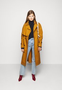Gul trenchcoat med dobbeltbrystet design, svarte knapper og brede revers. Båret over en svart polo-genser og kombinert med lyseblå jeans.