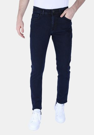 Man draagt slim-fit donkerblauwe jeans en witte sneakers, met één hand in de zak, staand tegen een effen lichte achtergrond.