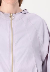 Lichtpaarse jas met een ritssluiting aan de voorkant, voorzien van een gouden rits, een capuchon en een textuurstof. Duidelijk zichtbare stiksels en naaddetails.