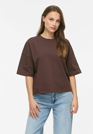 Femme aux longs cheveux bruns portant un chemisier ample à manches courtes marron foncé et un jean bleu clair, debout devant un fond uni.