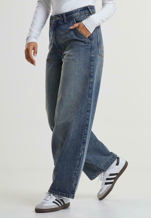 Personne portant un jean bleu large, une chemise blanche à manches longues et des baskets blanches à rayures noires, une main dans la poche, marchant.