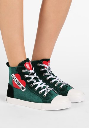 Grüne Samt-High-Top-Sneaker mit weißen Sohlen und Schnürsenkeln, roten Herz-Patches und "LOVE MOSCHINO" markierten Details auf Schnürsenkeln und Seiten.