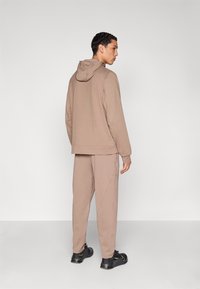 Καφέ φούτερ με κουκούλα και κορδόνι, με ομοιόμορφη υφή υφάσματος, σε συνδυασμό με ταιριαστά καφέ παντελόνια φόρμας με πλαϊνές τσέπες, φορεμένα με μαύρα αθλητικά παπούτσια.