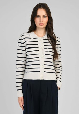 Femme portant un cardigan blanc à rayures horizontales noires et à gros boutons, associé à un pantalon noir plissé taille haute.