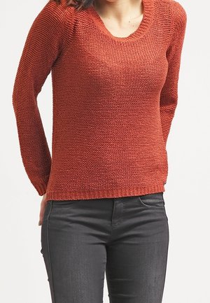 Kobieta w rdzawej, teksturowanej dzianinowej bluzie i ciemnoszarych dżinsach, stojąca z rękami za plecami.