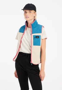 Izolovaný vest so modrým prešívaným vrchom a krémovým fleece spodkom, doplnený o ružový zips a zipsovú vrecko na hrudi.