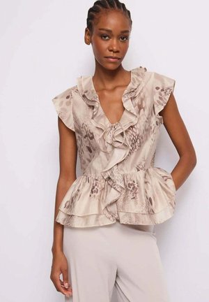 Donna che indossa una camicetta beige senza maniche con balze e una stampa animalier sottile, e pantaloni chiari, in piedi su uno sfondo bianco.