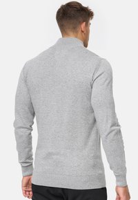 Mann iført en lys grå langermet genser, står med ryggen mot kameraet mot en hvit bakgrunn.