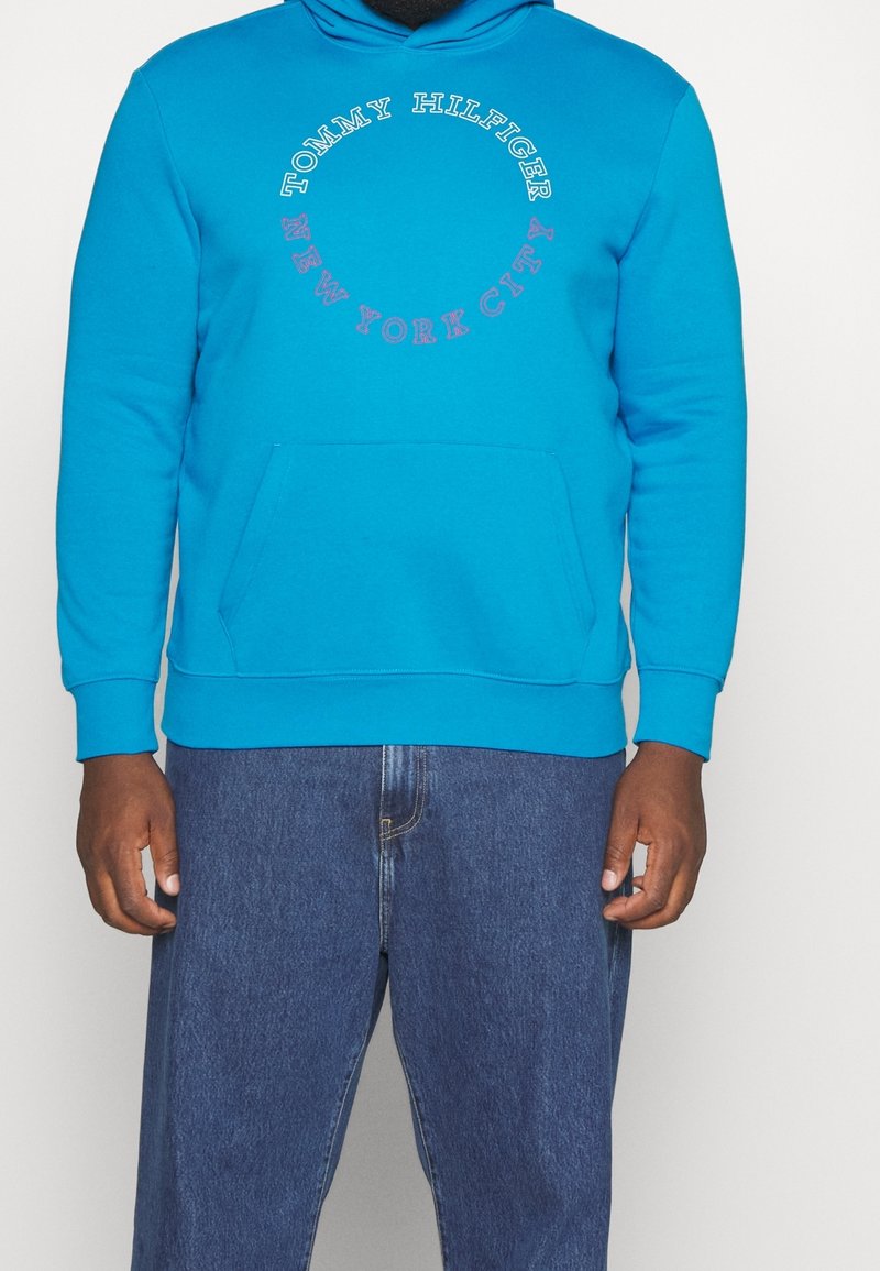 Homme portant un sweat à capuche Tommy Hilfiger bleu vif avec poche avant et cercle de texte indiquant "Tommy Hilfiger New York City" et un jean bleu.