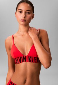 Bralette triangolare rossa con spalline sottili, dotata di un elastico con logo Calvin Klein nero. Texture liscia, design minimalista.