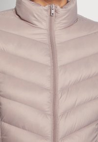 Ljusrosa puffjacka med hög krage, med en framzipper och ett chevron-quiltat mönster. Slät, lätt tygtextur.