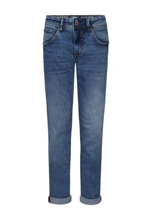 Personne portant un t-shirt noir, un jean bleu et des baskets blanches, debout devant un fond blanc uni.