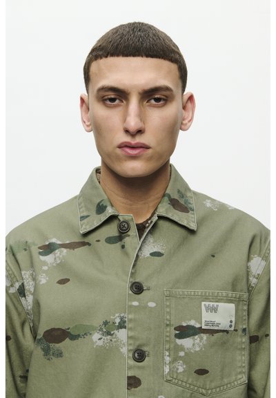 Chaqueta de algodón verde con patrón de camuflaje, que cuenta con un cuello, cierre de botones y un bolsillo en el pecho con una etiqueta de la marca.