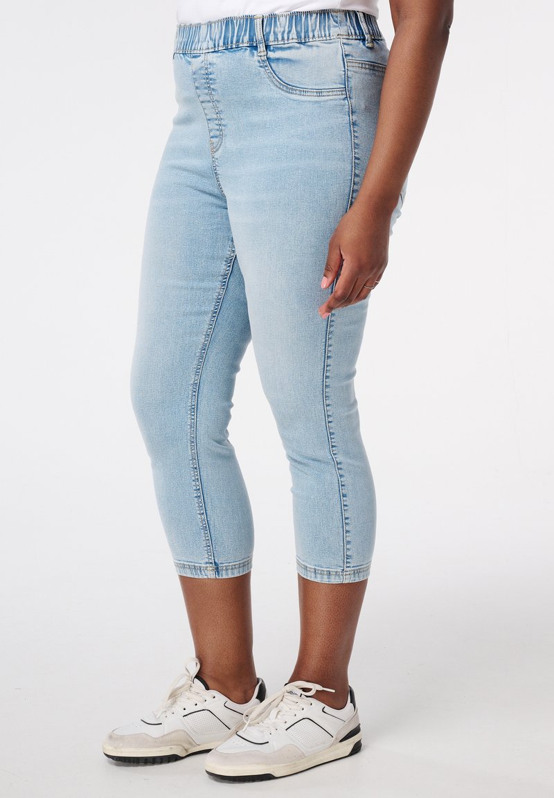 Jean capri en denim stretch bleu clair avec une taille élastique, présentant une coupe ajustée et un design classique à cinq poches. Porté avec des baskets blanches.