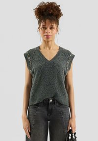 Woman with curly hair in a high bun wearing a sleeveless gray knit sweater with embellishments, black jeans, and holding a black handbag.