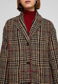 Cappotto a quadri nei colori beige, nero e rosso. Caratterizzato da un colletto a punte, chiusura con bottoni e due tasche frontali. Tessuto testurizzato.