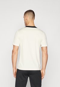 Uomo con capelli corti che indossa una camicia bianca a maniche corte con colletto nero, abbinata a pantaloni neri, in piedi davanti a uno sfondo semplice.