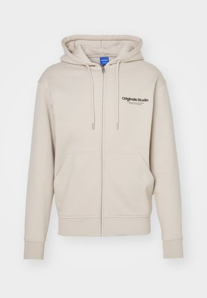 Beige zip-up hoodie with a drawstring hood, two front pockets, ribbed cuffs, and "Originals Studio" logo on the upper left. Smooth texture.