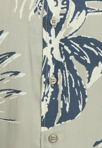 Chemise à boutons en tissu beige avec un motif floral bleu. Comprend quatre boutons, une texture lisse et des motifs contrastés sur la surface.