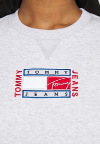 Primo piano di una persona che indossa una felpa grigia chiara con un logo rettangolare di Tommy Jeans in rosso, bianco, blu e nero sul petto.