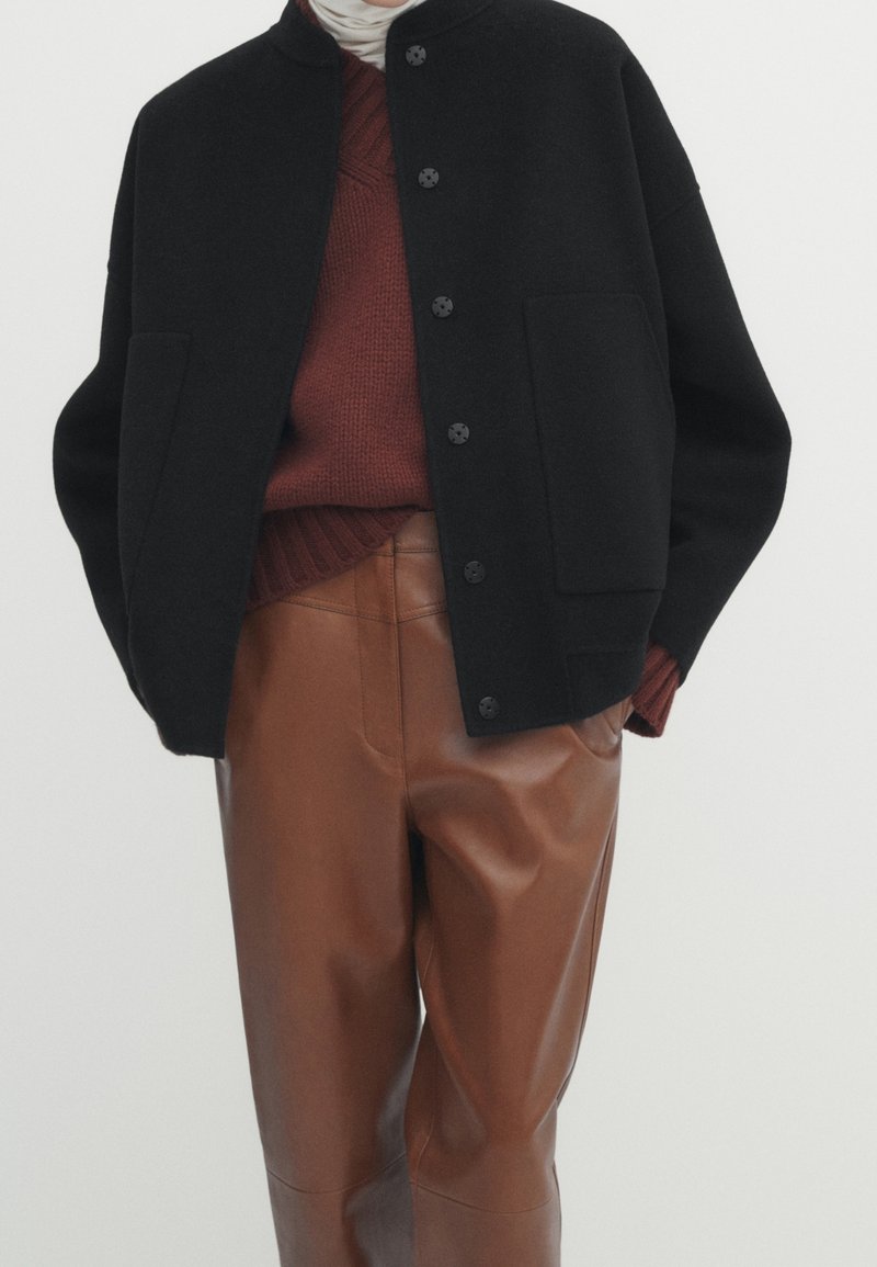 Veste noire en mélange de laine avec un col rond, des boutons à l'avant et deux poches, portée par-dessus un pull en tricot bordeaux et un pantalon en faux cuir marron.