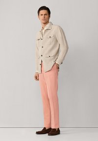 Mężczyzna stojący w beżowej kurtce z kieszeniami na klapach, białej koszuli, różowych spodniach i brązowych mokasynach na jednolitym jasnoszarym tle.