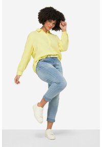 Camisa amarilla de botonadura con una tela texturizada, jeans azules remangados y zapatillas blancas. La camisa tiene un cuello y un corte holgado.