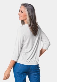 Weißes Oberteil mit Dreiviertelärmel, runder Ausschnitt und gerafftem Saum, kombiniert mit blauen Jeans. Glatte Textur, lässiges Design.