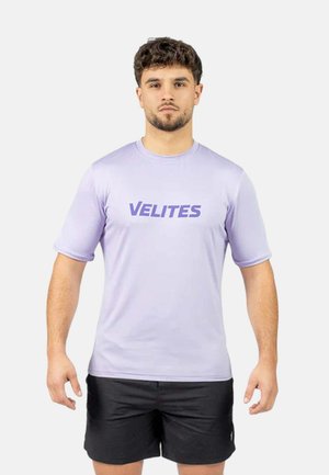 Joven con pelo corto y rizado, lleva una camiseta lila clara con el texto "VELITES" y pantalones cortos negros, de pie frente a un fondo liso.
