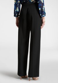 Pantaloni neri a gamba larga con una texture liscia e una vita, visti da dietro. Comprendono scarpe col tacco alte di colore marrone chiaro.