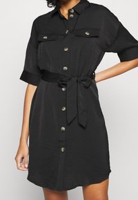 Black button-up dress with short sleeves, two front pockets, a tie belt, and a soft, textured fabric. Features round buttons and a relaxed fit.
