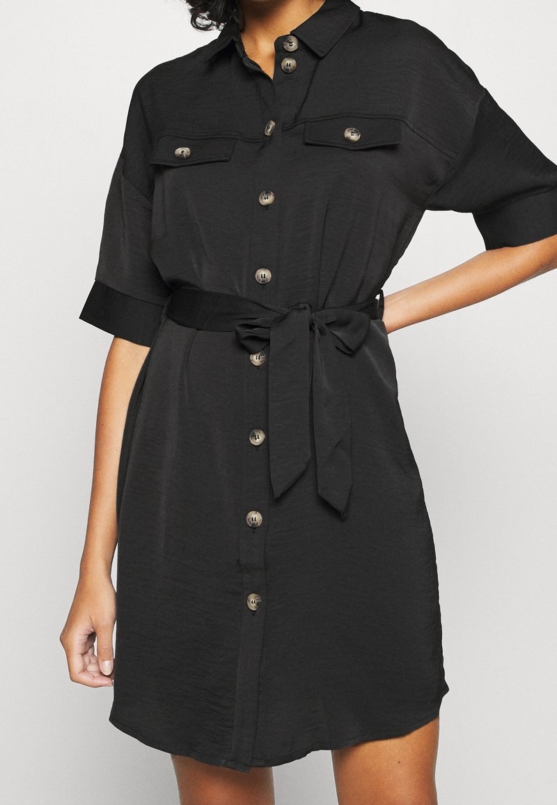 Black button-up dress with short sleeves, two front pockets, a tie belt, and a soft, textured fabric. Features round buttons and a relaxed fit.