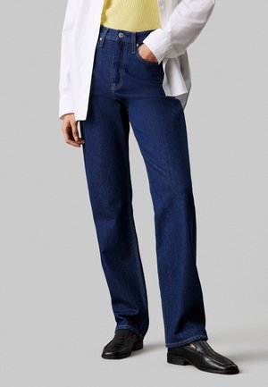 Jeans de mezclilla azul oscuro con corte recto, cintura alta y costuras en contraste. Combinados con una blusa amarilla clara y zapatos negros.