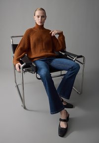 Rust-colored, ribbed knit turtleneck sweater, paired with flared blue denim jeans and black slingback shoes, seated in a metal chair.