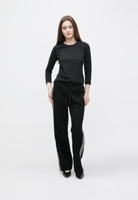 Crna majica dugih rukava uparena s crnim hlačama za trčanje s bočnim prugama, pojasom s vezicama i suženim krojem. Model nosi crne cipele.