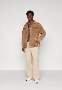 Bruine corduroy jas met twee borstzakken, gecombineerd met een effen bruin overhemd en lichtbeige wijde pijpen broek. Schoenen zijn lichtgekleurde sneakers.