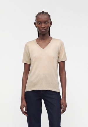 Femme aux cheveux tressés portant un haut beige à manches courtes en V et un jean foncé, debout devant un fond blanc uni.