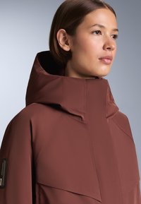 Barna vízálló dzseki magas gallérral és letisztult, minimalistikus dizájnnal. Matt felületű és varrott díszítésekkel rendelkezik.