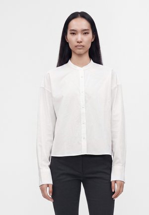 Femme aux longs cheveux foncés vêtue d’une chemise blanche boutonnée avec un col mandarin et un pantalon noir sur un fond clair uni.