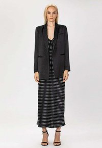 Blazer noir avec une texture satinée, porté sur une robe sans manches noire à pois. Des escarpins noirs à lanières complètent le look.