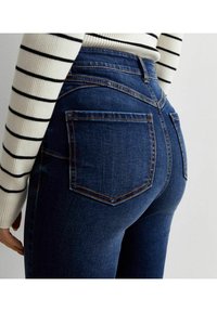 Jeans azul oscuro con un diseño ajustado, que cuentan con bolsillos traseros, costuras en contraste y una cintura alta. La modelo lleva una blusa de manga larga a rayas.