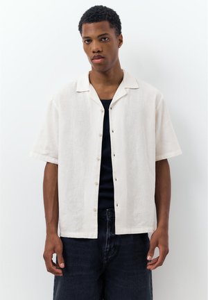 Giovane uomo con indosso una camicia bianca a maniche corte aperta sopra una maglietta nera e jeans scuri, su uno sfondo bianco semplice.