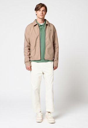 Joven de pie con chaqueta beige, camiseta polo verde, pantalones blancos y zapatillas blancas sobre un fondo blanco liso.