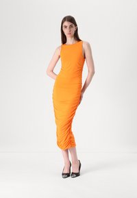 Robe midi orange avec des détails froncés, design sans manches et silhouette ajustée. Portée avec des escarpins noirs à bout pointu.