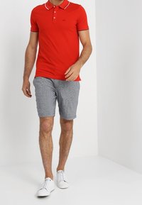 Man som går iklädd en röd kortärmad polo-tröja, grårutiga shorts och vita sneakers framför en enfärgad vit bakgrund.