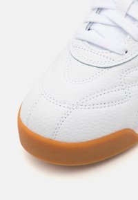 Hvide lædersneakers med tekstureret overflade, der har en tan gummisål og hvide snørebånd. Syning detalje langs tåområdet.
