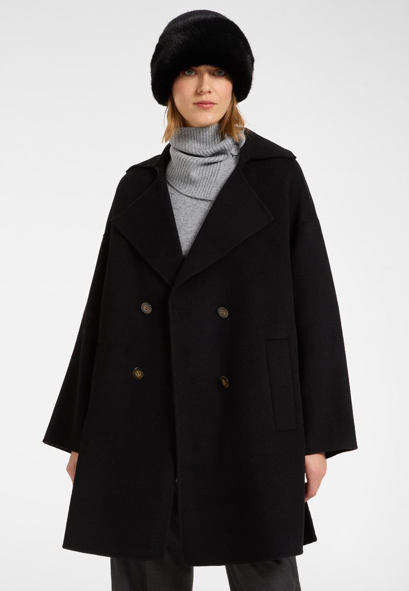 Abrigo negro de doble botonadura con grandes solapas, bolsillos delanteros y botones dorados. Combinado con un sombrero de piel negro y un jersey de cuello alto gris.