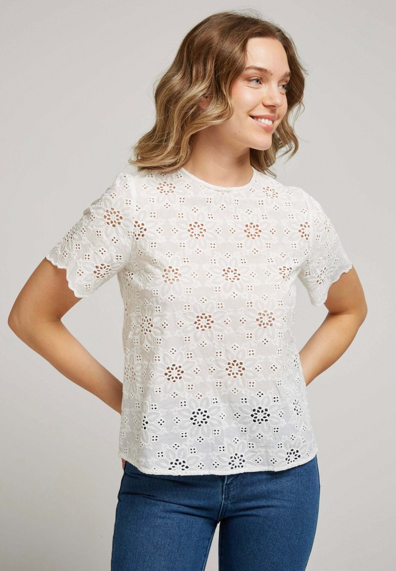 Blouse blanche à manches courtes brodée, ornée de motifs floraux et de découpes. Le tissu est léger et texturé.