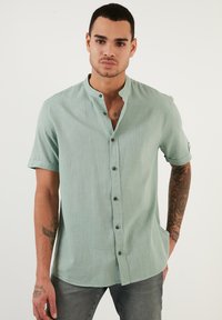 Camisa de manga corta verde claro con cuello mandarín, cierre de botones y una textura sutil. Presenta botones oscuros y un dobladillo curvado.